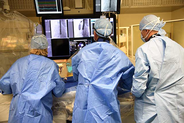 Photographie de l'équipe en train de pratiquer de dos, avec vue sur les écrans de contrôle lors de l'opération de correction d’une communication interatriale Sinus venosus par cathétérisme interventionnel