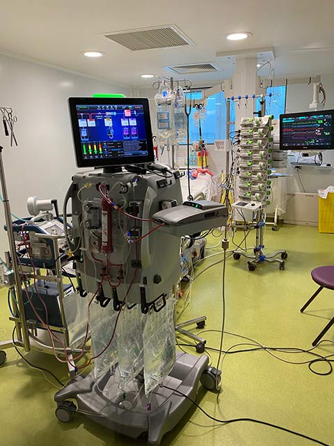 Un chirurgien en tenue stérile verte opère un patient allongé sur une table d'opération recouverte de champs chirurgicaux bleus dans un bloc opératoire équipé de divers appareils médicaux.